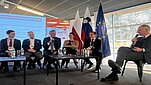 Sześć osób siedzi i rozmawia w pokoju konferencyjnym. Dwójka z nich trzyma mikrofony. Za nimi flagi: Unii Europejskiej, Słowenii i Polski